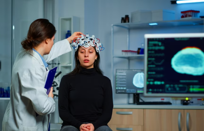 A woman sitting in front of a monitor while another person is putting on a hat.